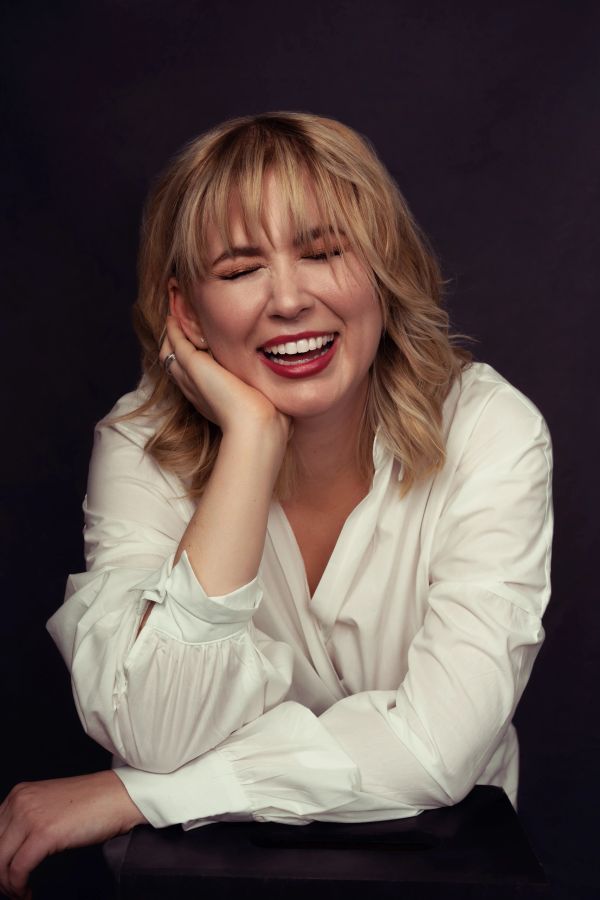 Blonde woman in white shirt laughing with eyes closed against dark background.