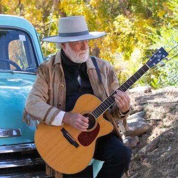 Elderly man in a wide-brimmed hat playing acoustic guitar outdoors.