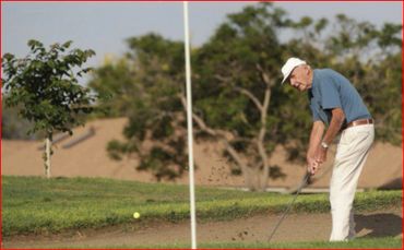 Elderly man playing golf, hitting ball from sand bunker.