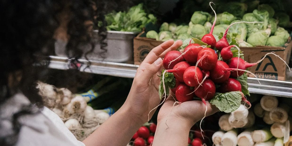 Woman's hands holding a bunch of radishes in the grocery store. Also on the shelf are other veggies