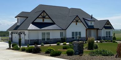 Front view of home with steep pitch black roof white siding lower stone and natural wood eves.