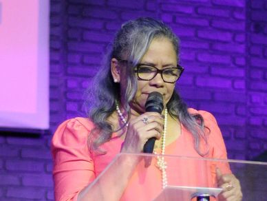 Woman speaking into a microphone at a podium with a purple brick background.