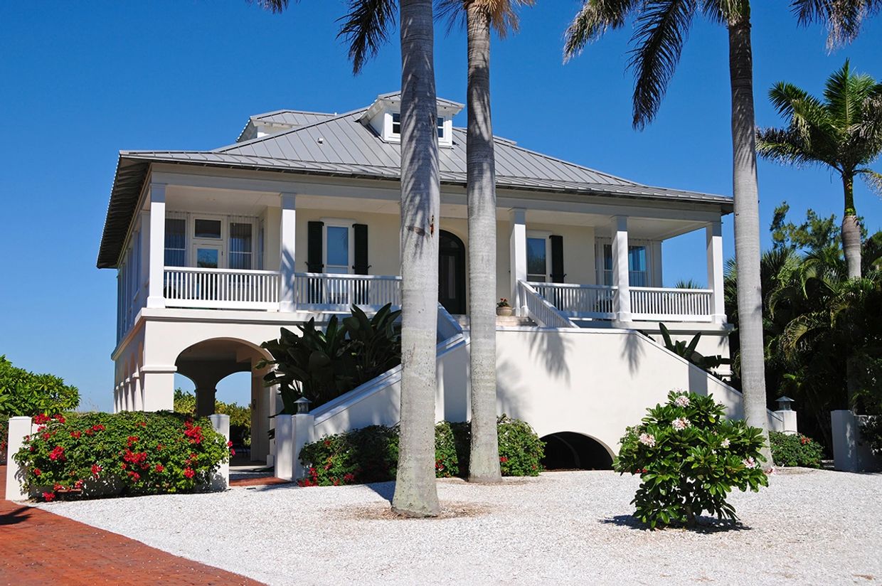 White coastal house with palm trees and a gravel driveway under a clear blue sky.