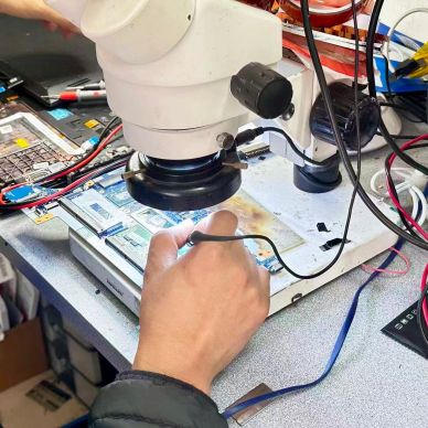 A person uses a microscope to repair a small electronic device on a cluttered workbench.