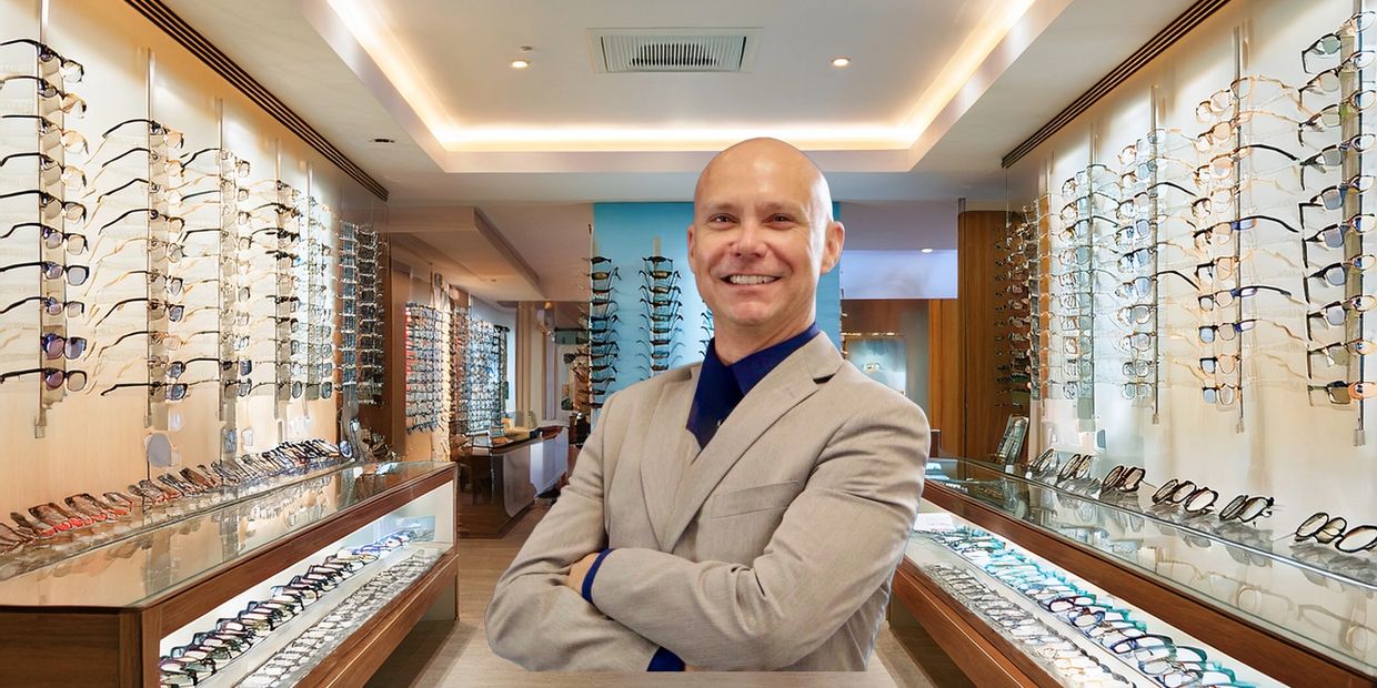 A smiling man in a beige suit stands in an eyeglass store.