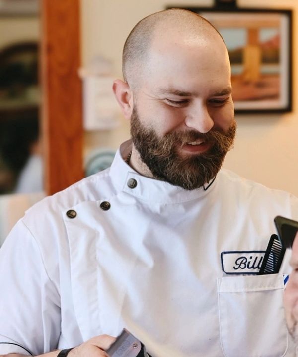 Smiling barber wearing a white coat with the name Bill embroidered.