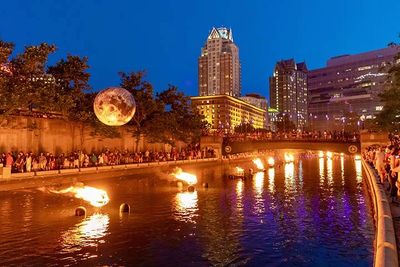 Nighttime river scene with fire fountains and a glowing moon art installation.