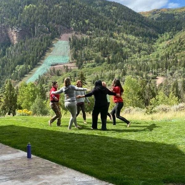 Six people holding hands in a circle outdoors with a mountain backdrop.