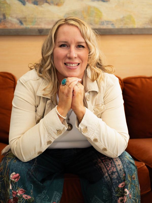 Smiling woman with blonde hair sitting on a couch, hands clasped.