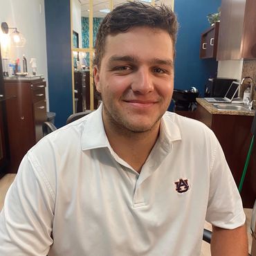 A young man smiling in a cozy indoor setting wearing a white Auburn University shirt.