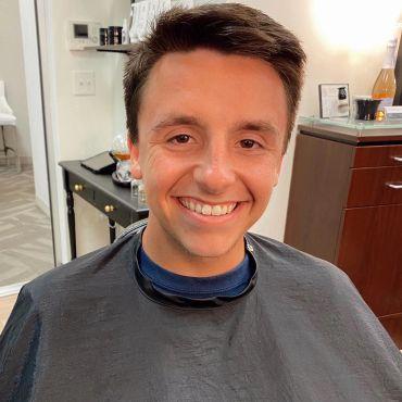 A young man smiling, wearing a barber cape in a salon.