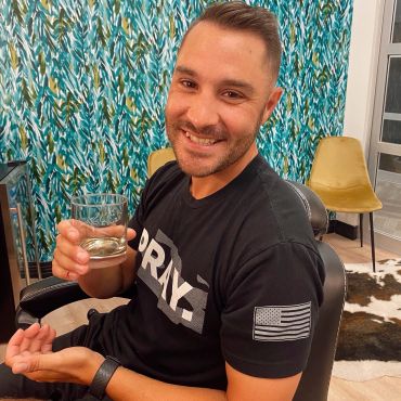 A man smiling while holding a glass of water indoors.