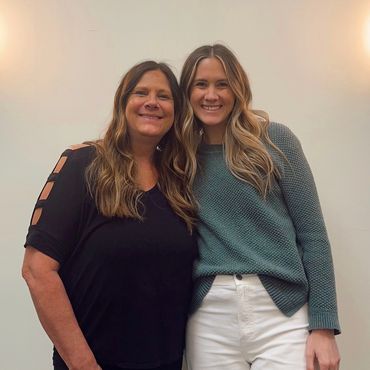 Two women smiling and posing together against a plain wall.
