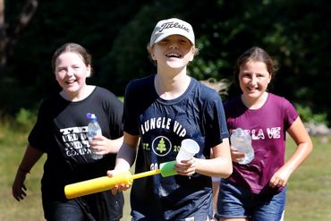 Three girls smiling outdoors, holding water bottles and a toy bat.