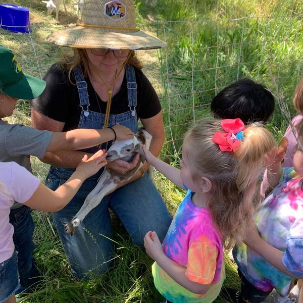 Children petting a small goat held by a woman in a hat and overalls.