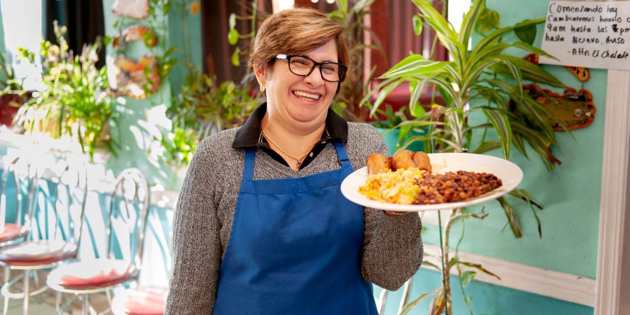 Marta, smiling, wearing a blue apron and holding a plate of El Salvadorian food.