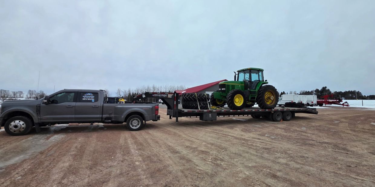 Pickup truck towing a large flatbed trailer with a green tractor and extra tires.