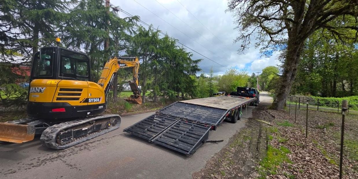 Yellow excavator beside a flatbed trailer on a rural road.