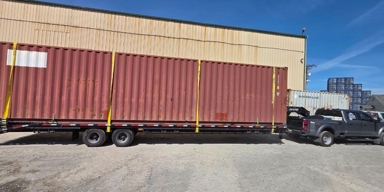 A pickup truck towing a flatbed trailer with a large red shipping container secured by yellow straps.