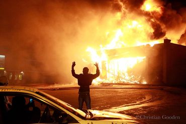 A person raises hands in front of a large building fire at night.
