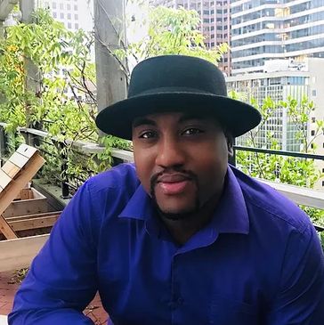 Man in a black hat and blue shirt smiling on a balcony.