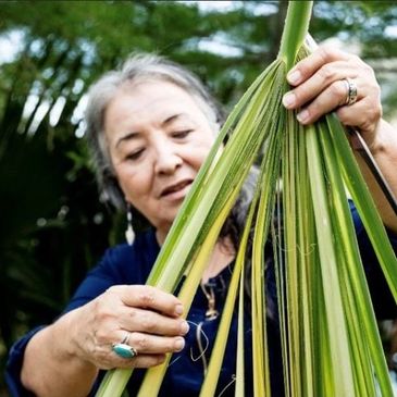 An elderly woman weaving green palm leaves outdoors.