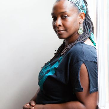 A woman with dreadlocks wearing a headband and earrings, standing by a window.