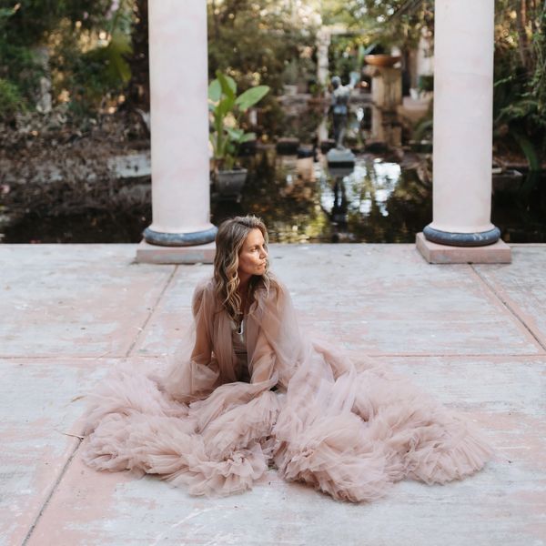 Woman in a flowing beige gown sits gracefully on a stone patio near columns and a reflective pond.