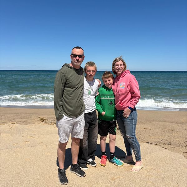 Family of four posing on a sandy beach with ocean and clear sky.
