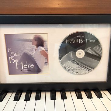 Framed CD and album cover of 'I'll Still Be Here' by Katey Ann on a piano.