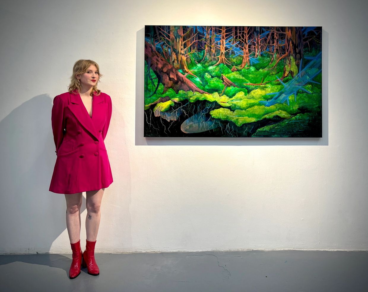 Woman in a bright pink blazer dress stands beside a vibrant forest-themed painting in a gallery.