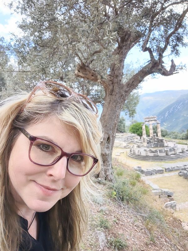 Woman with glasses taking a selfie near ancient ruins and a large tree.