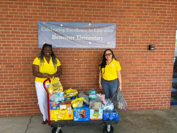 Two women in yellow shirts with a cart full of supplies in front of a school banner.