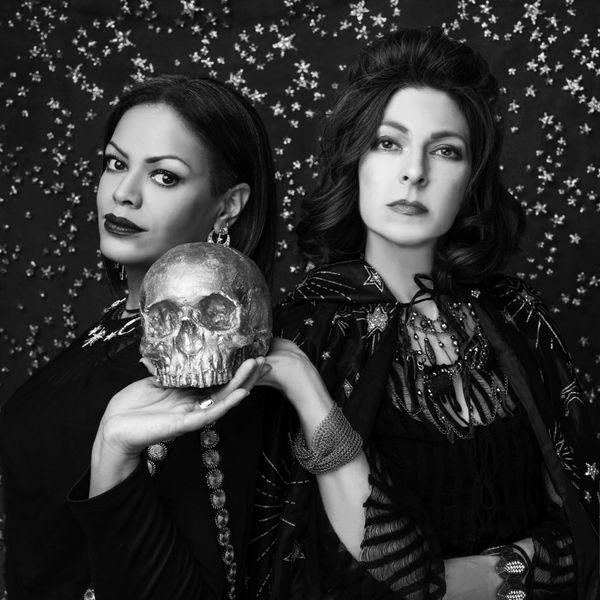 Two women in dark attire hold a metallic skull against a starry backdrop.