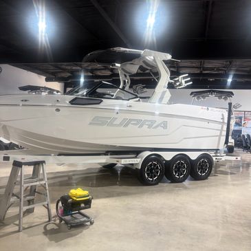 A sleek white Supra boat on a triple-axle trailer inside a showroom.