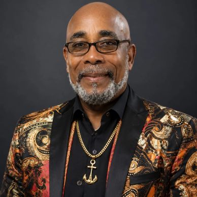 Smiling man wearing glasses and a patterned jacket with an anchor necklace.