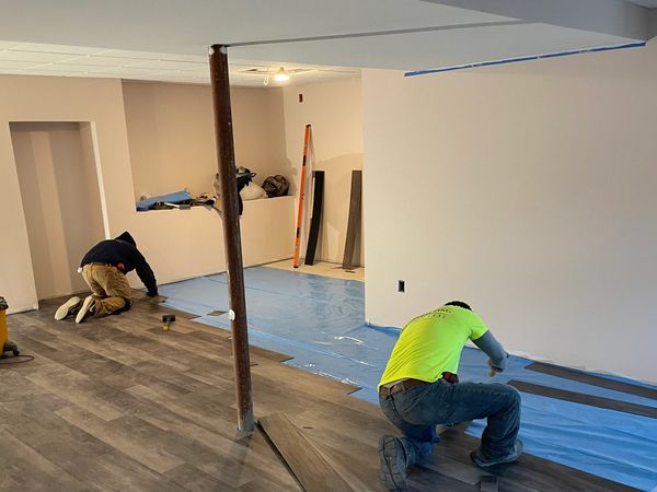 Two workers installing gray wood-look flooring.