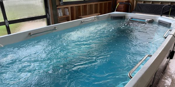 Indoor swim spa with clear blue water and metal handrails.