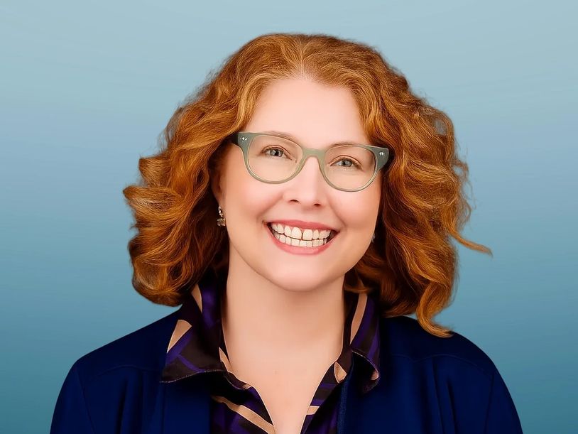 Smiling red-haired woman with glasses in a blue outfit against a blue background.