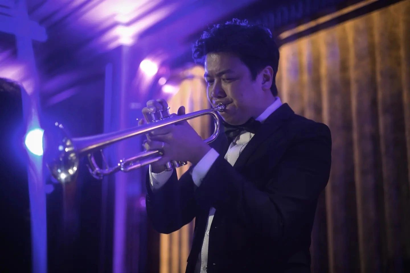 Man playing trumpet in formal attire under purple lighting.