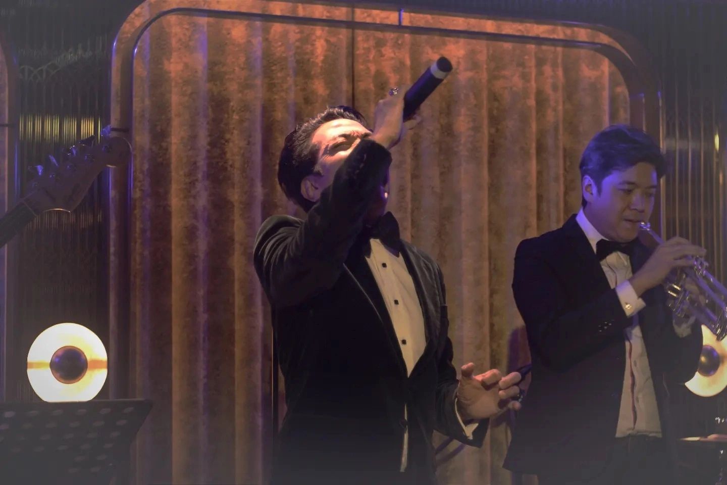 Two men in tuxedos performing on stage, one singing, one playing trumpet.