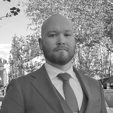 Man in a suit standing outdoors with trees and buildings in the background.