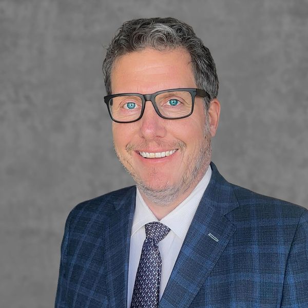 Smiling man in glasses and a blue checkered suit jacket against a gray background.