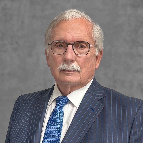Elderly man with glasses, white hair, and mustache in a pinstripe suit and patterned blue tie.