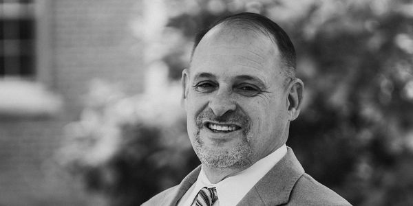 Smiling man in a suit standing outdoors with a blurred background.