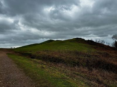 A cloudy sky over a green hill with a bench on the path.