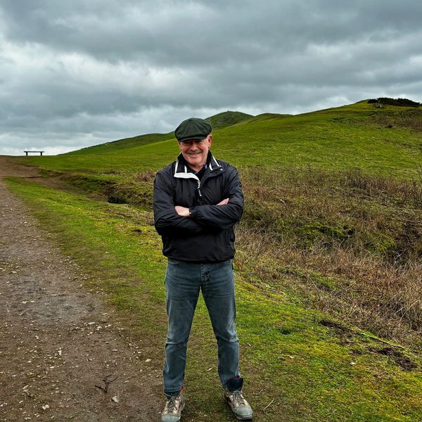 A man in a flat cap smiling on a green hill under a cloudy sky.