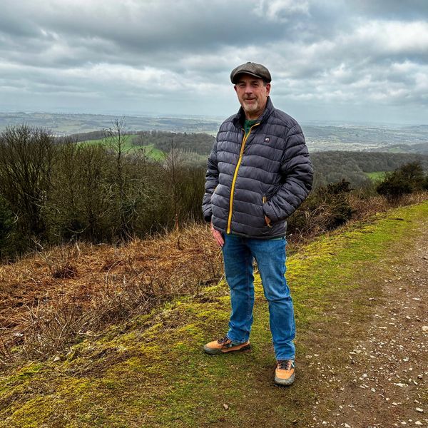 Man in outdoor jacket and cap standing on a hiking trail with a scenic landscape behind.