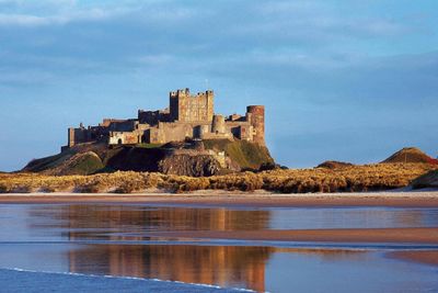 Historic castle on a hill reflected in calm water under a blue sky.