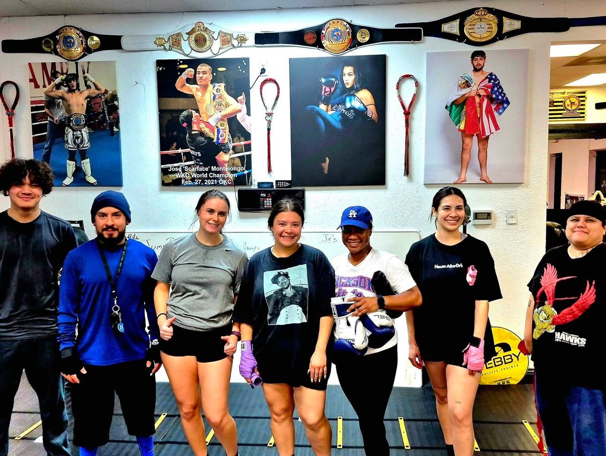 Group of seven people posing inside a martial arts gym with championship belts and boxing posters.